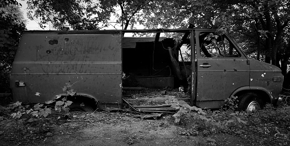 A monochrome image of a van that has been abandoned in a forest.