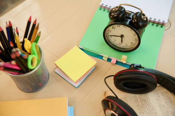 istock Top view black alarm clock on open notepad with school supplies and stationery on blurred foreground. Back to school 1415435708