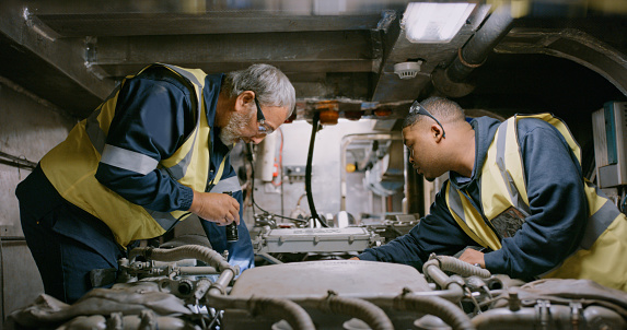 Mechanics at work, fixing and concentrating on engine an in repair. Two men wearing overall uniforms on the job in a large workshop. At the auto service, male colleagues examine a motor in a garage.