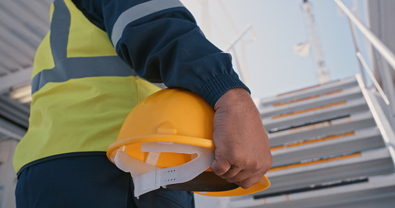 Closeup of male construction, builder or engineers hands holding a protective hardhat helmet on site. Industrial building worker or manager at workplace with yellow safety hat.