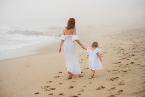 a young mother wearing a romantic white dress and her daughter wearing a matching dress are walking along the ocean barefoot.
