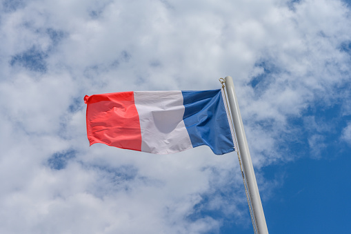 French flag waving in the wind. It is a beautiful sunny summer day, with blue sky and white clouds in the background.
