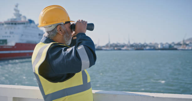 um técnico de navios olhando para a distância no oceano usando binóculos para verificar o nível do mar e a segurança. engenheiro de navios trabalhando no convés em um porto de importação e exportação - industrial ship dock worker engineer harbor - fotografias e filmes do acervo