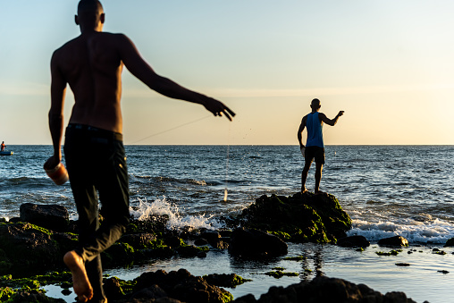 Salvador, Bahia, Brazil - December 11, 2021: Two fishermen fishing from above the rocks of Rio Vermelho beach in Salvador, Bahia.