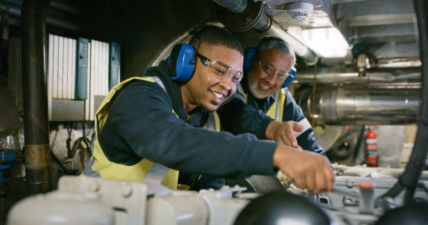 mecánico experto senior que entrena a un aprendiz en una planta de fabricación. hombres sonrientes en uniforme que trabajan en la reparación de motores en la fábrica con gafas de seguridad. mecánico profesional en desarrollo - aprendiz fotografías e imágenes de stock