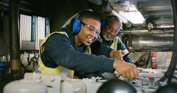 Two smiling men, in uniform and working on engine repair in factory wearing safety glasses. Senior expert mechanic coaching a young guy in a manufacturing plant. Professional mechanic in development