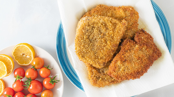 Crispy pan fried breaded pork chops served with lemon and fresh tomatoes close up on a plate on light grey background, flat lay