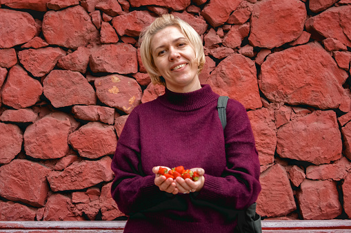 young smiling blonde woman in a red sweater against the background of a red stone wall holds large ripe strawberries in her palms