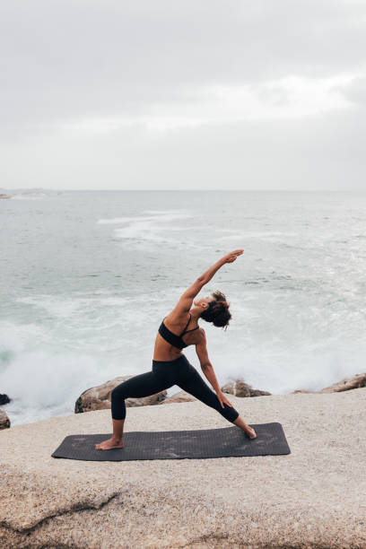 istock Female stretching her body while standing on a mat against ocean 1415460045