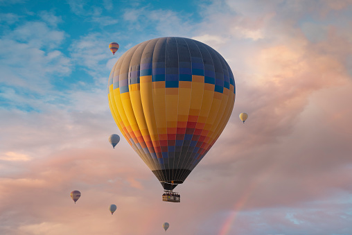Creative abstract colorful travel, tourism aerial transportation and freedom concept: 3D render illustration of color rainbow hot air balloon with gondola basket outdoors in the blue sky with clouds