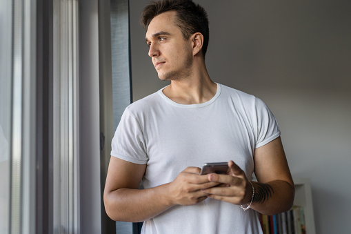 One man young adult caucasian male standing by the window at home wear white shirt using mobile phone to send sms messages or browsing internet via social network app application install copy space