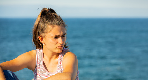 young girl in a tank top with a pensive look and the sea in the background with copy space in a bright sunset