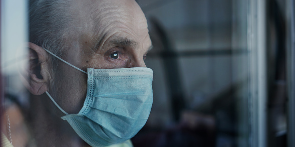 Side close view of older man in surgical facemask looking with sorrow through glass of window indoor