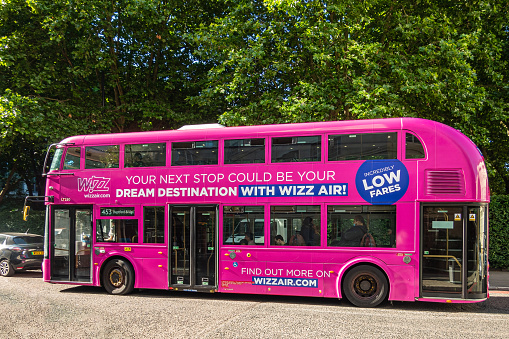 London, UK - July 5, 2022: Pink double decker public bus with ad on side for Wizz Air on Albany street, Green foliage in back.