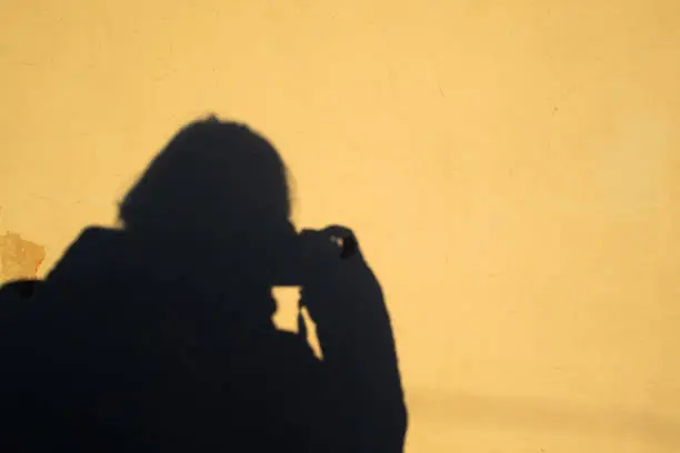 Photo of Selfie of a female photographer in the form of a human shadow with a camera on a yellow old shabby wall background. Copy space