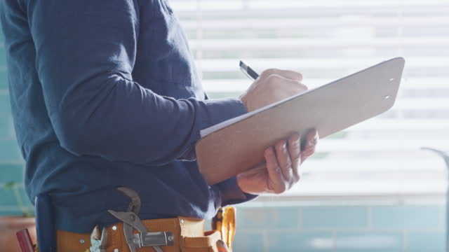 Plumber and maintenance worker writing on clipboard, checking fixed household appliance and monitoring repair work. Closeup hands of handyman or electrician preparing an invoice after finishing a job