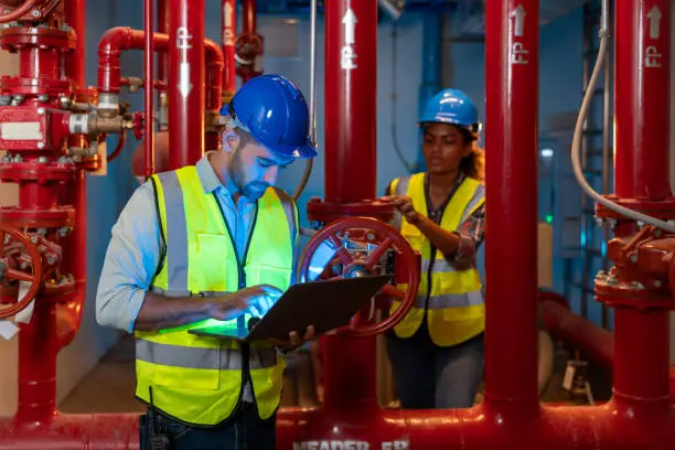 Engineer use laptop to inspection pipeline and valve of service water supply in fire riser room. Multi-Ethnic Blue-collar worker and technician testing fire suppression system. Fire protection concept