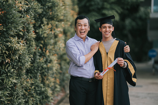 proud Asian Malay father and son in college graduation gown looking at camera smiling after receiving the certificate diploma