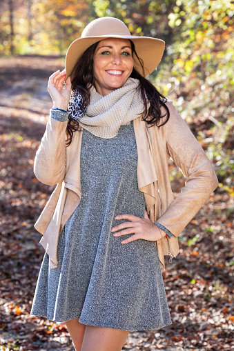 Charming woman enjoying sunny autumn day in the forest. Trees in the background. Wearing grey mini dress, leather jacket, ankle boots and wide-brimmed hat.