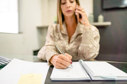 Close-up of a young businesswoman writing down notes and talking with a colleague on a phone while working at her desk in an office