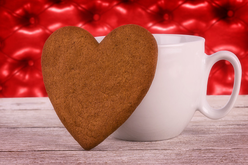 Heart shaped hand made cookie and white coffee mug on red leather capitone in background. Valentines day concept.
