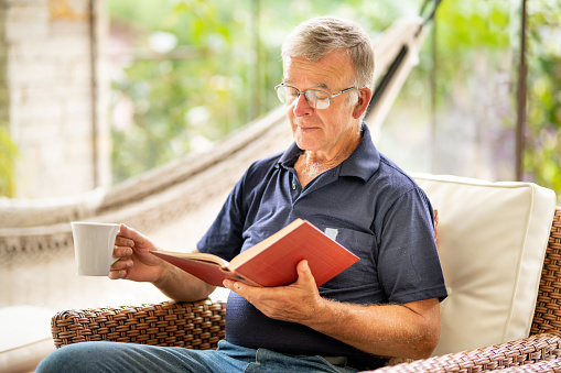 Elderly man, book and coffee on sofa with smile, reading or relax in retirement in home living room. Senior person, literature and happy with tea cup for knowledge, thinking and drink on lounge couch
