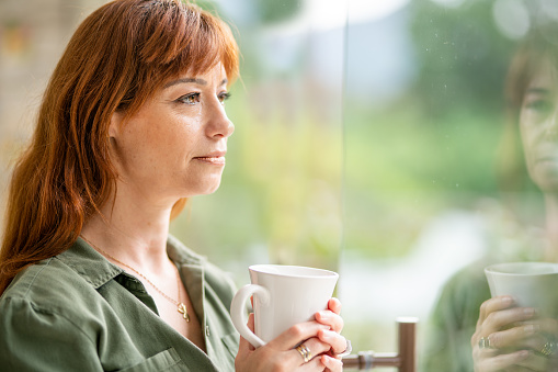 Woman looking lost in thought while drinking a coffee and looking out through a window at home