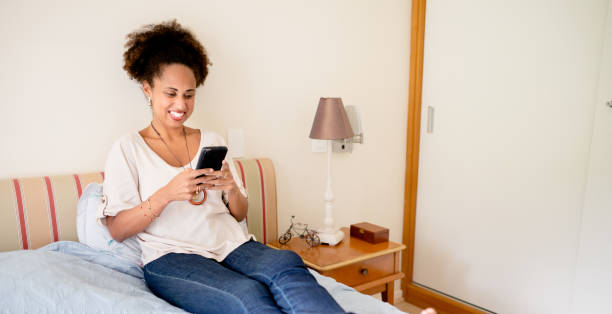 istock Woman smiling and sending a text on her phone while sitting on her bed 1415491424