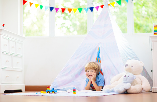 Kids room with teepee tent. Children play in white sunny bedroom. Cozy light interior for kid nursery or playroom. Little boy with toy blocks and dinosaur playing on wooden floor. Home decoration.