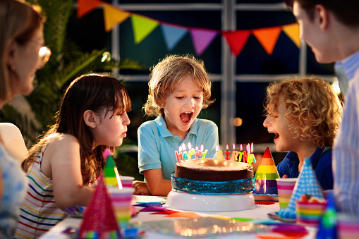 Kids birthday party. Child blowing candles on cake and opening presents. Pastel rainbow theme celebration. Family celebrating at home. Boy opening gifts, eating cakes. Sweets for children.