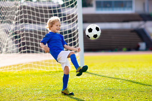 Kids play football on outdoor stadium field. Children score a goal during soccer game. Little boy kicking ball. School sports club. Training for young player.