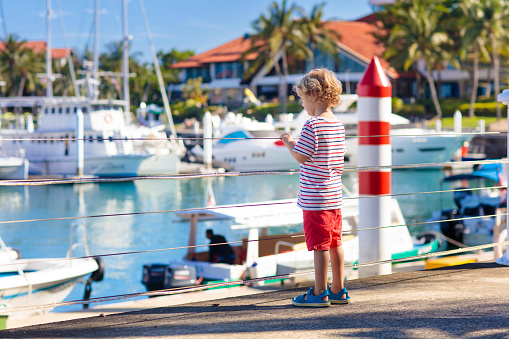 Child watching yacht and boat in harbor. Yachting sport for family with kids. Little boy walking in tropical resort on summer vacation. Nautical fashion for children. Holiday at the sea.