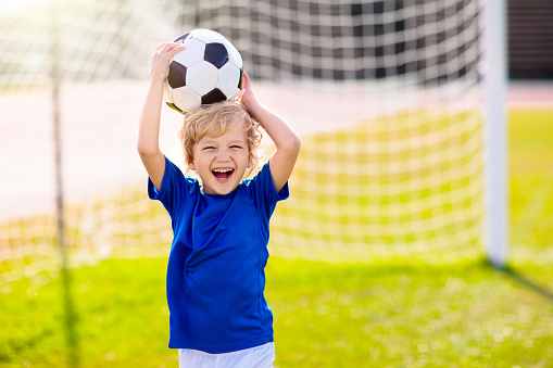 Kids play football on outdoor stadium field. Children score a goal during soccer game. Little boy kicking ball. School sports club. Training for young player.