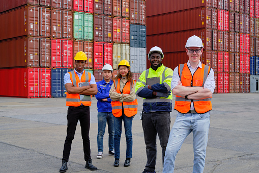 Portrait of a group of multiracial workers team in safety uniforms, arms crossed and looking at camera at logistics dock with many stacks of containers, shipping goods, cargo transportation industry.