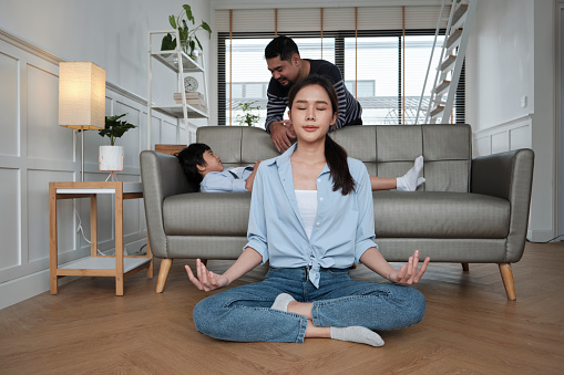 Young Asian Thai mother sits on living room floor, meditates, and practices yoga for health and wellness when husband and son tease together on sofa, happy domestic home lifestyle on family weekend.
