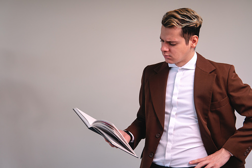 A blonde Caucasian man dressed in a brown jacket and white shirt with his hand on his waist reading a book on a white background. Teacher reading in a class. Worried lawyer with documents.