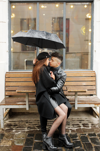 Young couple sitting near a cafe on a bench under an umbrella in rainy weather. Concept of love, romance and passion. November 2, 2019, Lviv, Ukraine.
