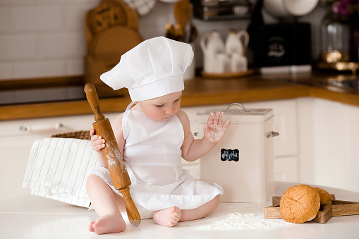Lifestyle. A little boy of 6 months in a cook's apron is sitting on the kitchen table with a rolling pin, a whisk and fresh bread.