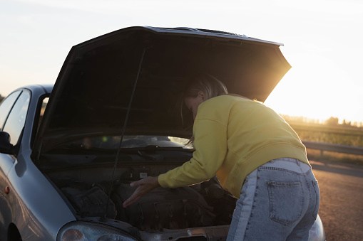 girl with blond hair in casual clothes stands with an open hood at sunset and looks at what has broken. A car breakdown on the road. There is a place for an inscription on the left. High quality photo