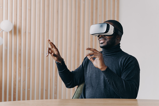 Joyful African man in VR headset glasses playing favorite 3D game while sits at desk at home office smiling and waving with hands up and down touching air. Innovative technology concept