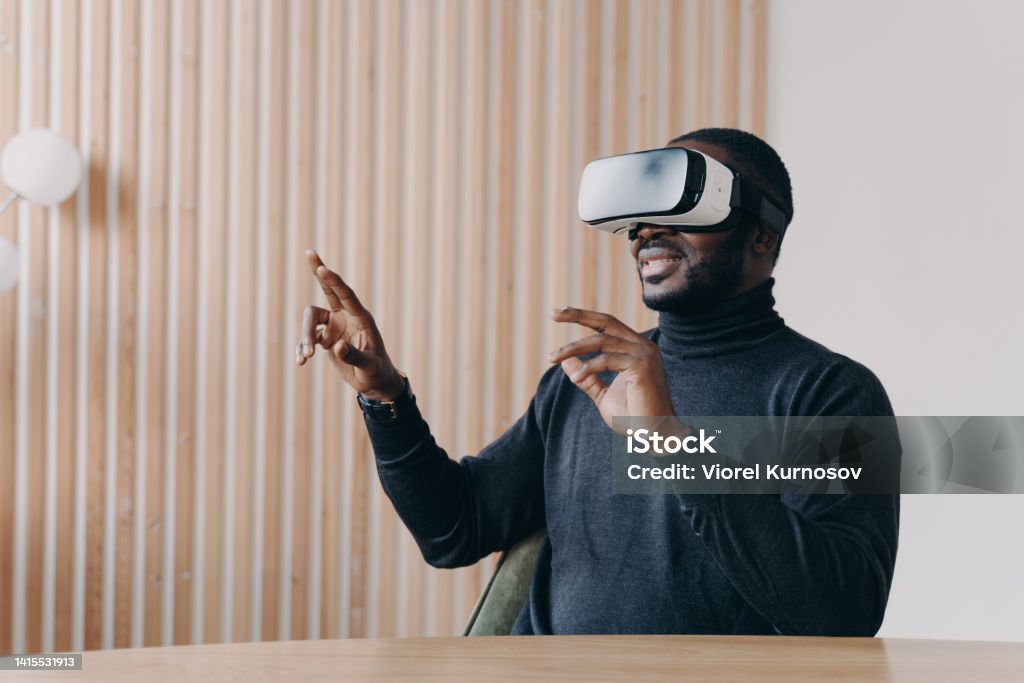 Young joyful African man office worker in VR headset glasses playing favorite 3D game Joyful African man in VR headset glasses playing favorite 3D game while sits at desk at home office smiling and waving with hands up and down touching air. Innovative technology concept Virtual Reality Stock Photo