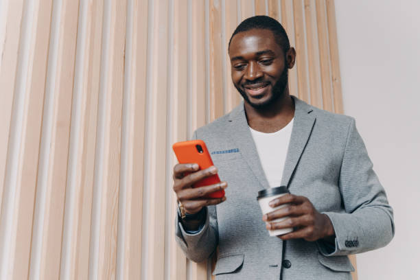 istock Enjoying coffee break at work. Young positive african man office worker in suit using mobile phone 1415531914