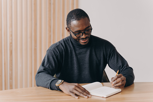 Young african american man entrepreneur in glasses taking notes in agenda while sitting at desk at home or office,planning workday, writing down some fresh ideas or work schedule