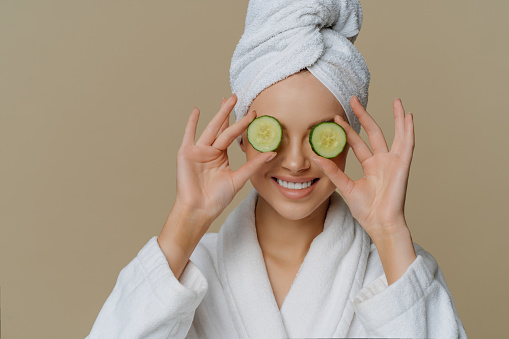 Indoor shot of pleased woman covers eyes with cucumber slices nourishes skin smiles happily dressed in bath dressing gown wrapped towel on head stands indoor. Beauty spa cosmetology concept.