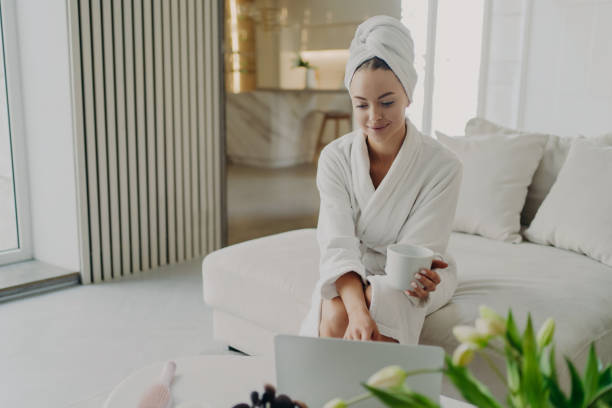 istock Young woman in white bathrobe working remotely on laptop while sitting on sofa in modern living room 1415534952