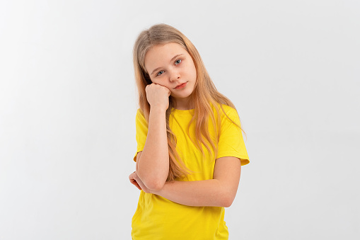 Cute teen girl looks bored or upset, sulks with head on fist, stands disappointed against in blank yellow tshirt over white background