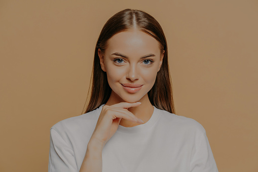 Lovely young European woman with makeup healthy skin touches gently jawline looks directly at camera has long straigh hair natural beauty dressed in casual sweater isolated over brown background