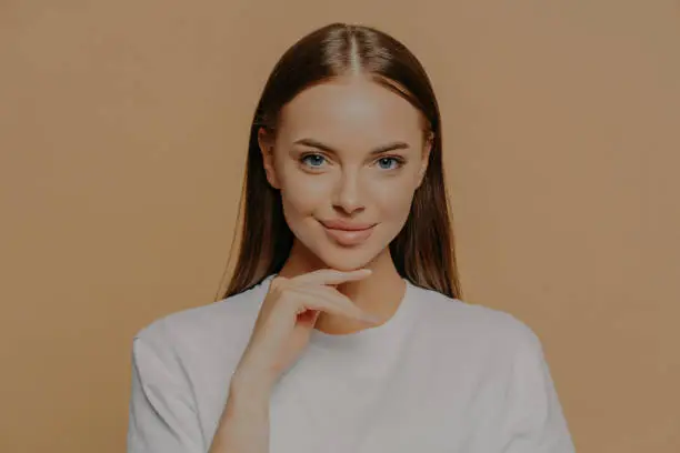Lovely young European woman with makeup healthy skin touches gently jawline looks directly at camera has long straigh hair natural beauty dressed in casual sweater isolated over brown background