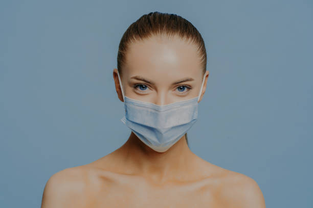 istock Young serious female model looks directly at camera stands bare shoulders indoor wears medical mask to protect coronavirus spread isolated over blue background. Health care and sickness concept 1415536132