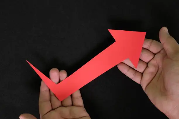 Human hands holding red upward arrow in dark black background. Business and economic recovery from crisis concept.